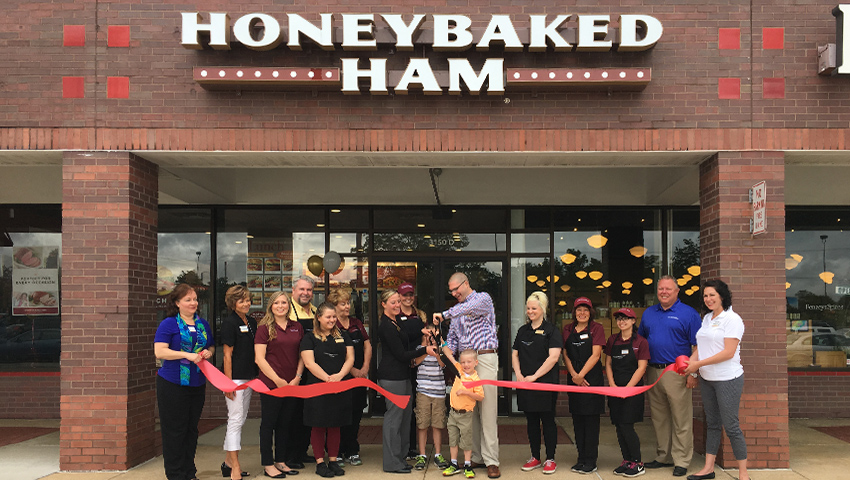 Ribbon-cutting at HoneyBaked Ham store-front with large group.