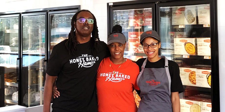 Franchisee Rosa Ramirez standing alongside with two of her employees in front of HoneyBaked fridges filled with retail products.