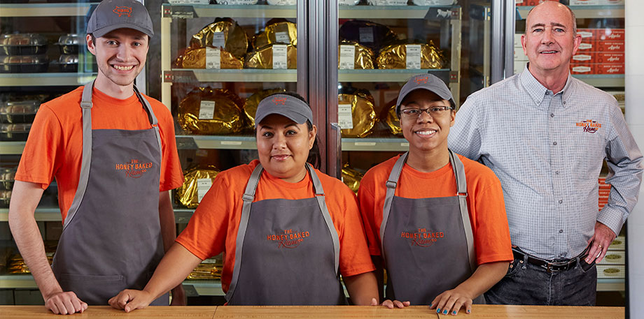 Three Honey Baked Ham employees with their franchisee store-owner standing behind the counter in front of retail products in refrigerators.
