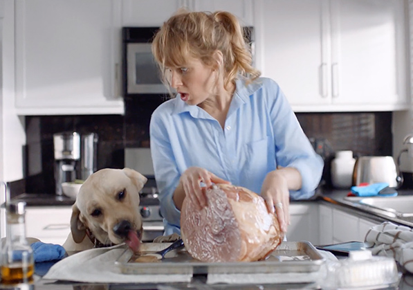 Blonde woman attempting to cook her ham in her kitchen but her lab dog is liking the honey off of the sheet pan the ham is placed on.