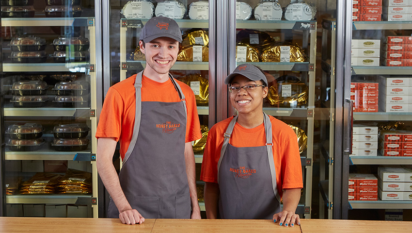 Two young people in HoneyBaked uniforms smiling.