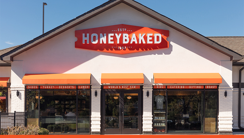 Exterior image of white Honey Baked Ham franchise store with orange brand sign and awnings