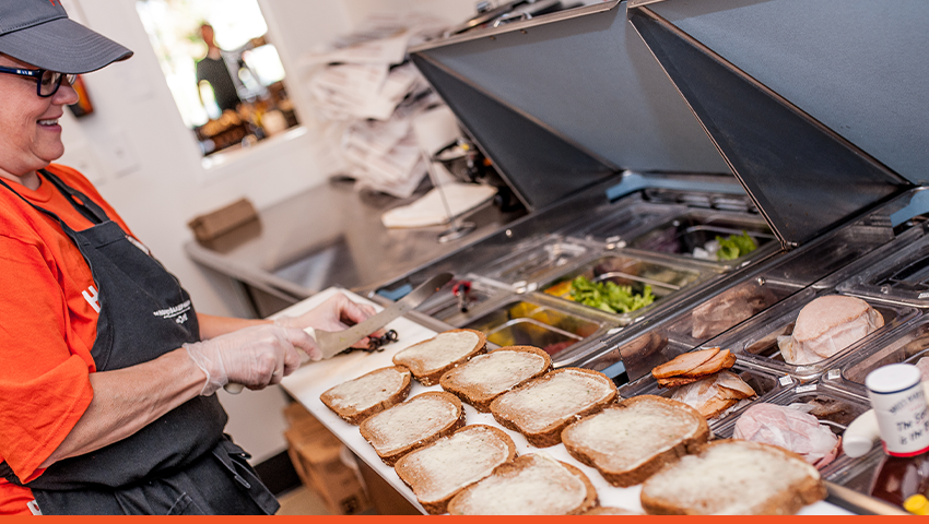 A HoneyBaked franchise employee makes five sandwiches at a restaurant prep station.