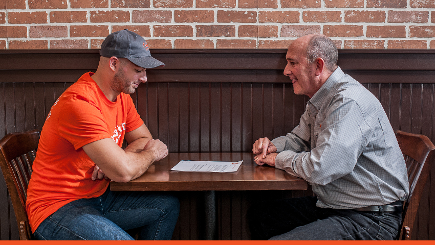 A male employee and a male customer sit together at a table in a HoneyBaked restaurant.